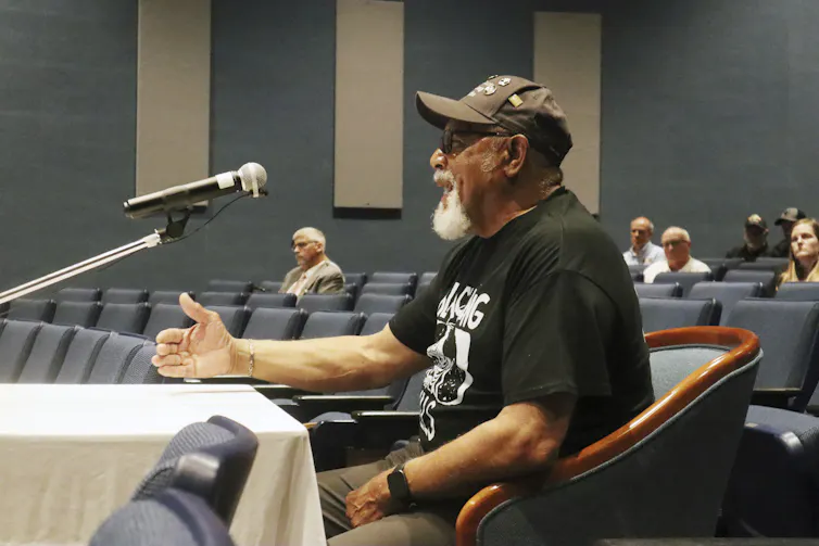 A man sits at a table and speaks into a microphone.