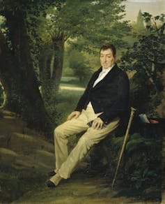 A seated, brown-haired man in a suit jacket and khaki-colored pants sits in a glen with his cane beside him.