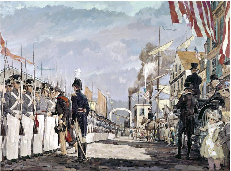 A line of men in uniform stand in formation on a street as onlookers stand nearby.