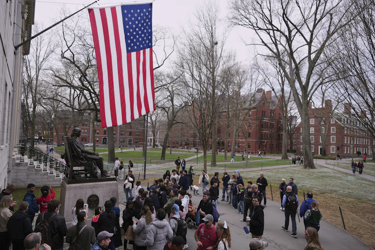 People gather round a statue of a seated man outdoors on a college campus, below a large U.S. flag.