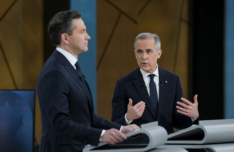 Un hombre con un cabello gris corto en el escenario de la gesticulación mientras hablaba mientras miraba el cabello oscuro, Zuric, recto de antemano.