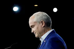 A bald man in a blue suit jacket photographed in silhouette looks down.
