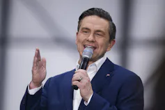 A dark-haired man in a blue suit gestures as he speaks into a microphone.