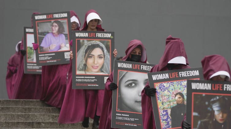 Las mujeres vestidas con vestidos para sustancias manuales rojos llevan fotos de mujeres asesinadas en Irán.