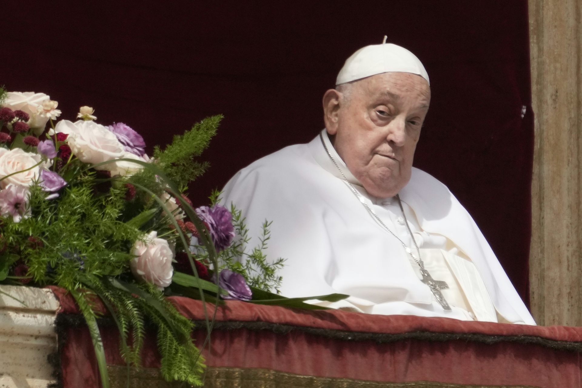 An elderly man in white robes sits next to a large bouquet of flowers.