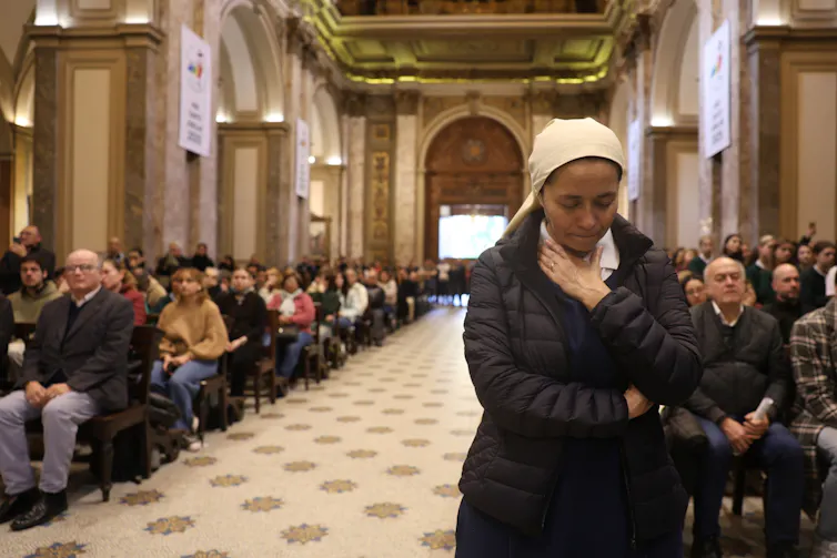 A woman standing with her head bowed, surrounded by other seated mourners.