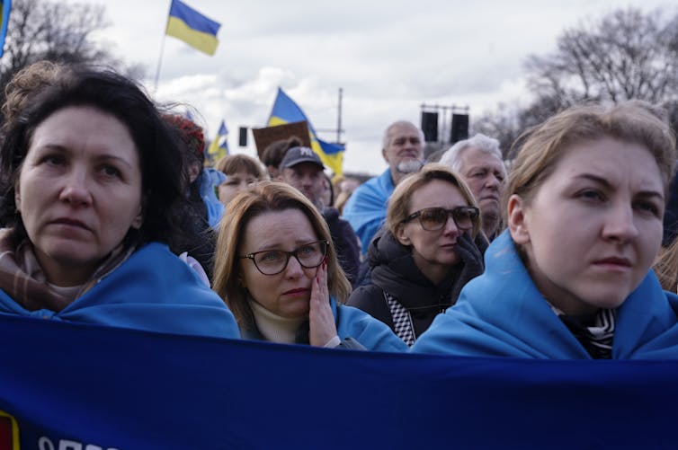 three women draped in ukrainian flags, one wipes away tears