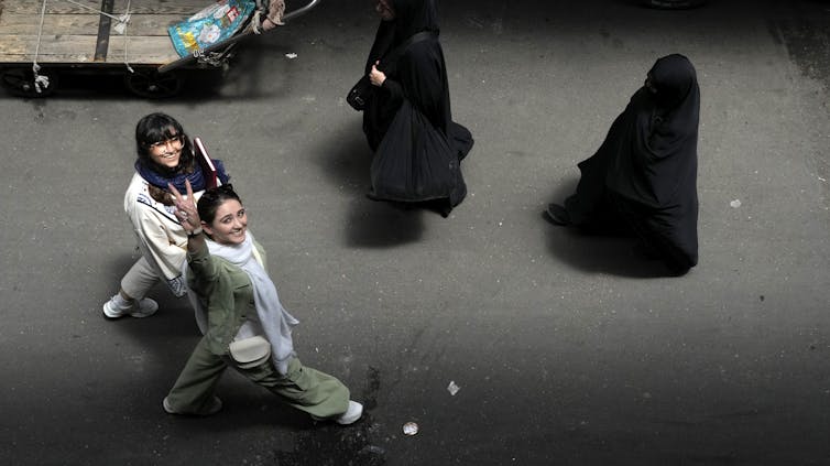 La mujer sonríe y levanta una señal de victoria mientras caminaba junto a la segunda mujer sonriente. Dos cortinas pasan junto a ellos.