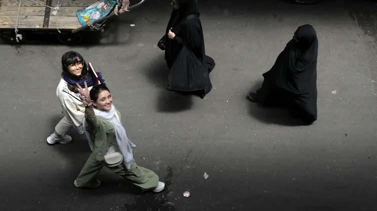 A woman smiles and raises a victory sign while walking next to another smiling woman. Two veiled women walk past them.