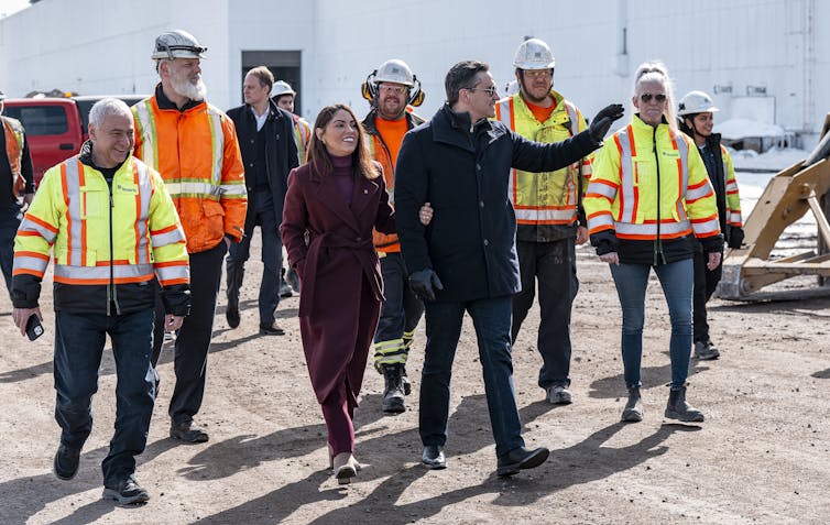 A dark-haired man and woman walk outdoors among manufacturing workers wearing safety vests.