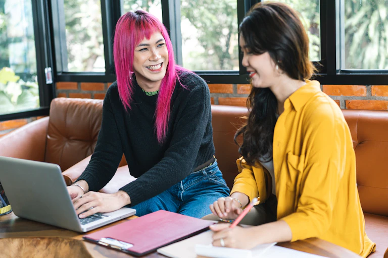 Smiling trans woman with pink hair speaks to another woman on a couch.