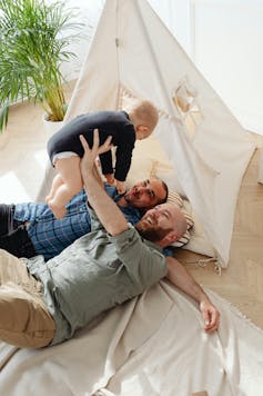 Two smiling men lying down while one holds their baby above them.