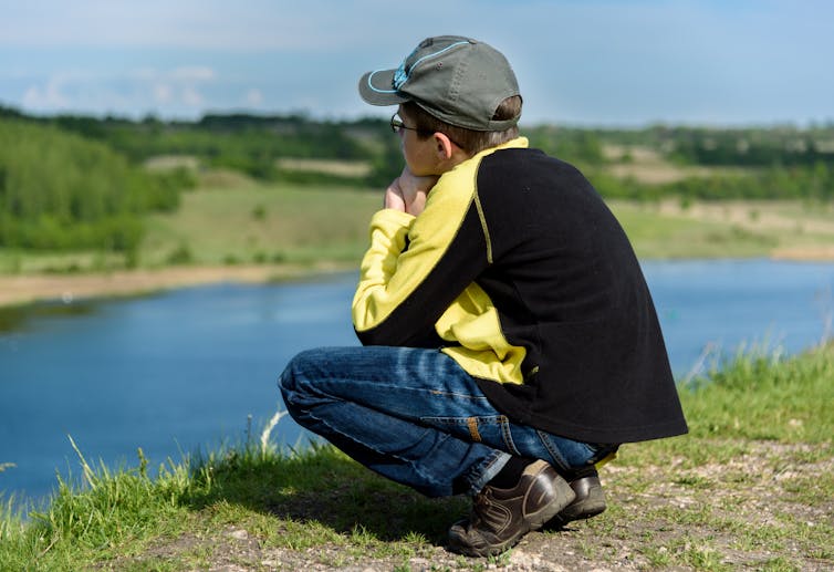 Teen boy crouches on river bank looking wistful.