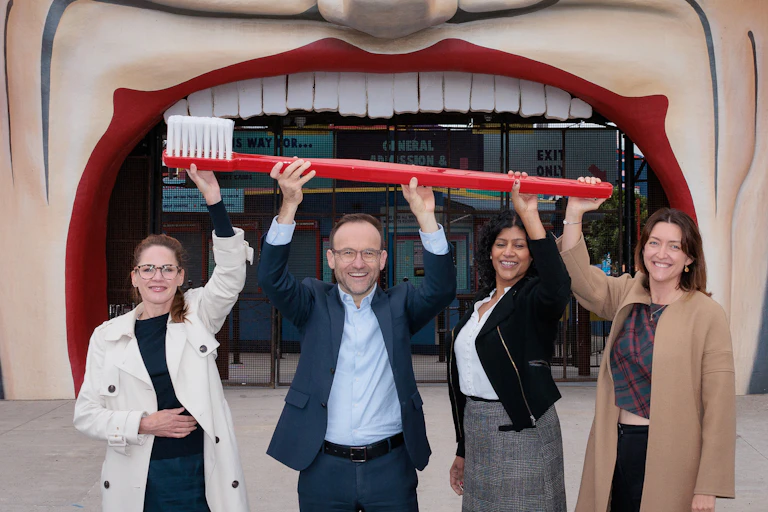 Three women and one man standing before a giant cartoon mouth holding a gigantic toothbrush