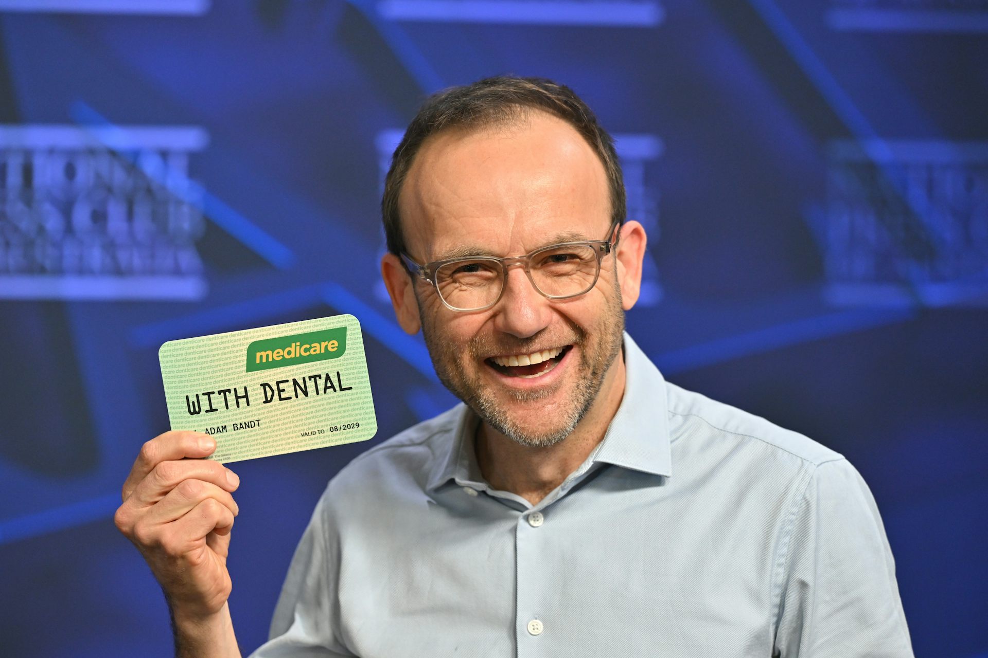 A laughing Adam Bandt wearing an open necked shirt and glasses and holding an oversized medicare card