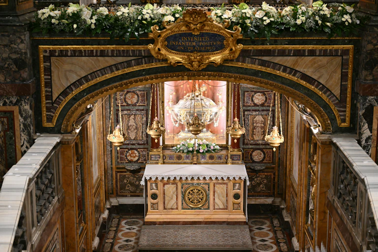 A precious wooden relic set inside an ornately decorated room, with a row of white flowers on a parapet above it.