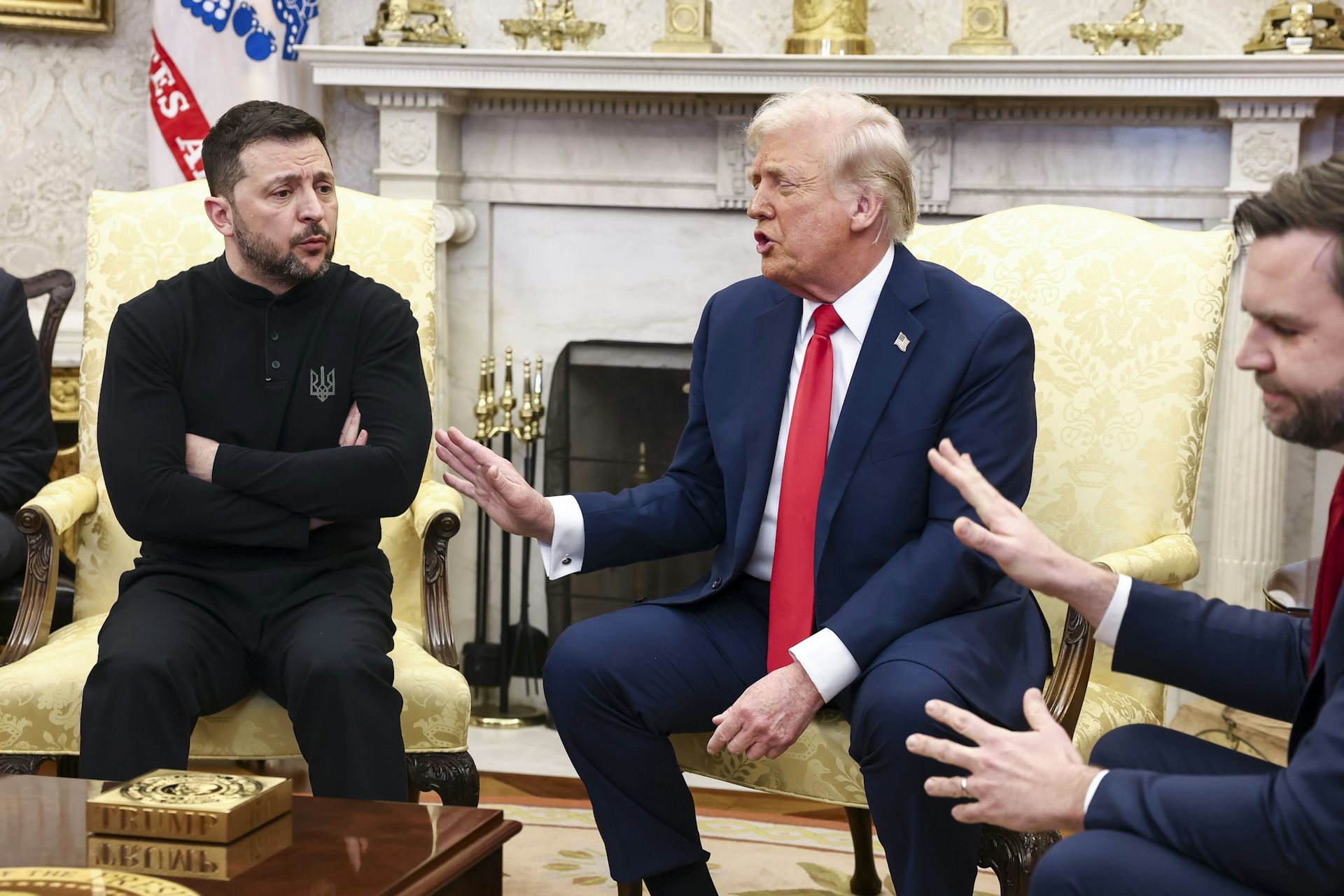 Ukraine's president, Volodymyr Zelensky, sits with his arms folded as US president Donald Trump and US vice-president JD Vance gesture at him.