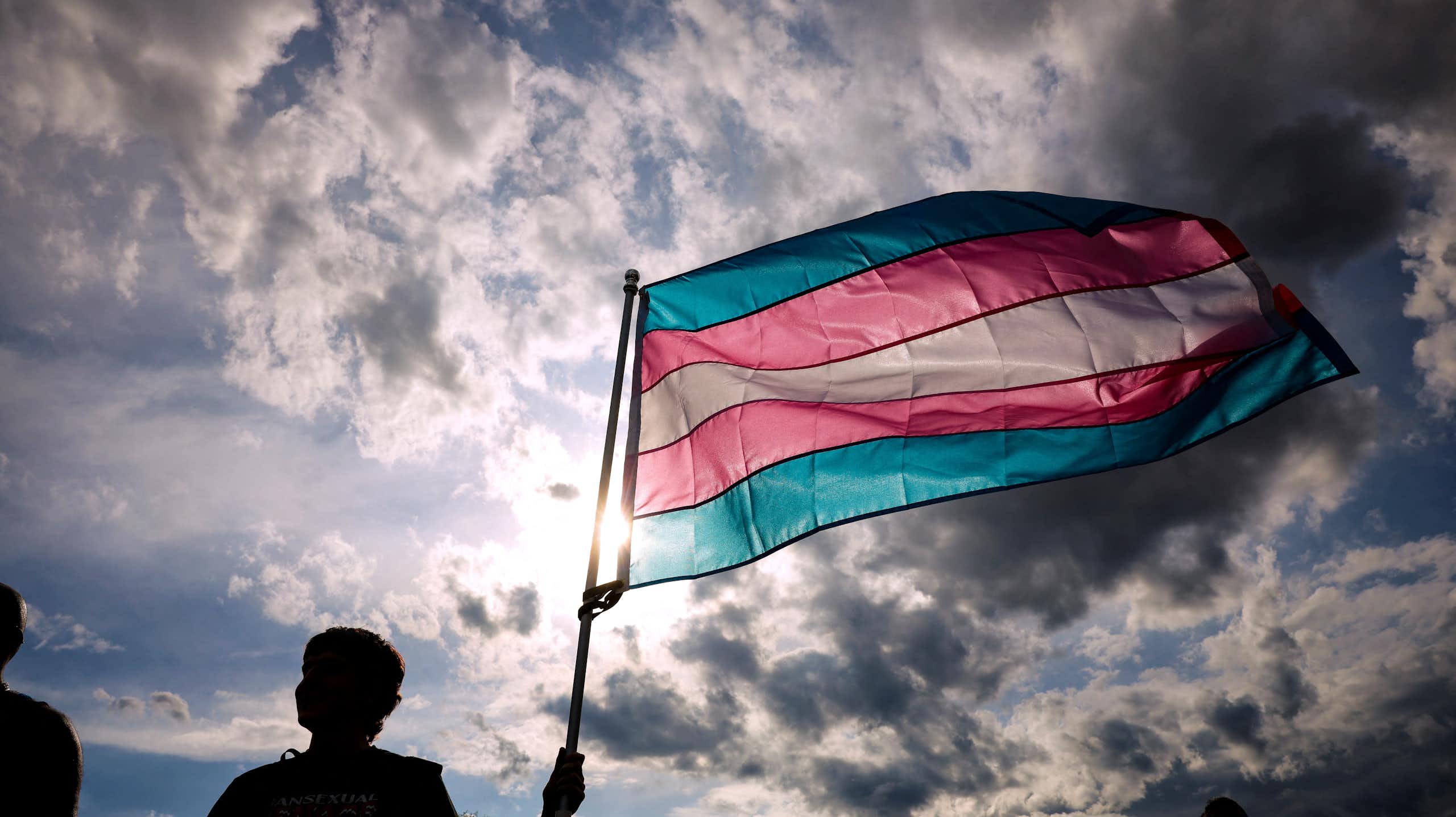 A person holds up a flag with horizontal stripes in light blue, pink and white underneath a cloudy sky with the sun shining through.