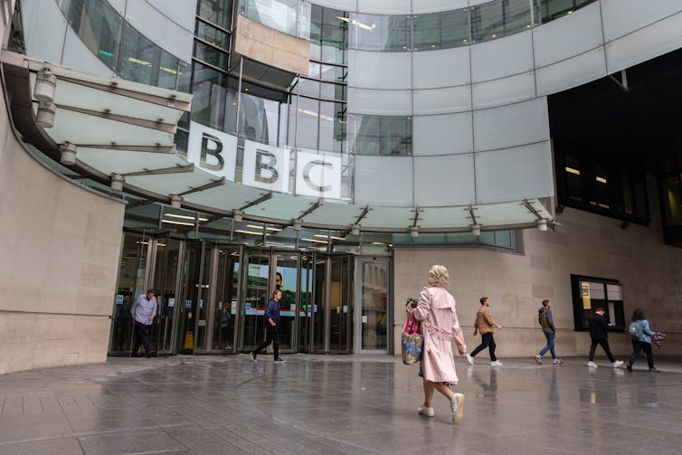 A woman in a light pink trenchcoat walks into the front of the BBC broadcasting house headquarters