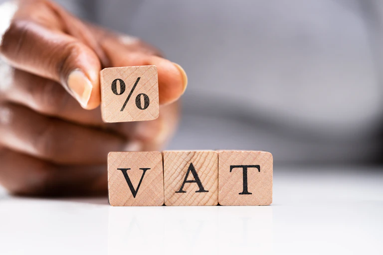 Fingers holding a wooden block bearing a percentage sign, placing the block on others that spell out VAT