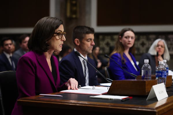 A woman with dark hair and a purple blazer sits at a table with other people dressed formally seated nearby her.