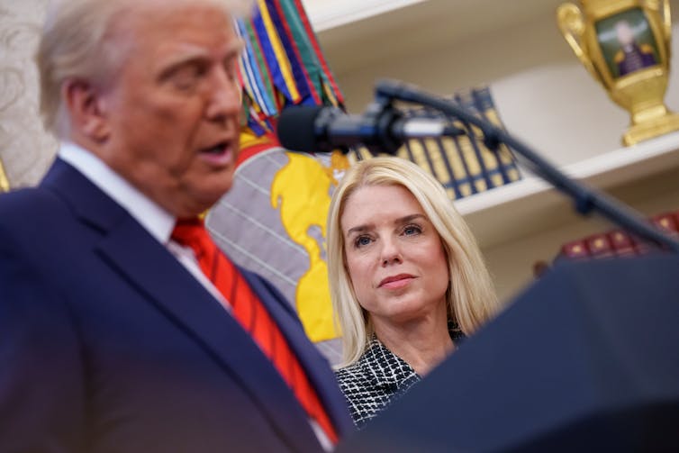 An older man with a blue suit speaks into a microphone while a woman with blonde hair looks at him.