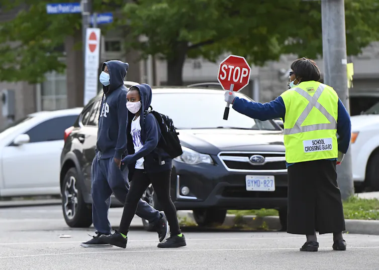 Two Black children crossing at a crosswalk as a Black crossing guard helps guide them across the street holding a stop sign.