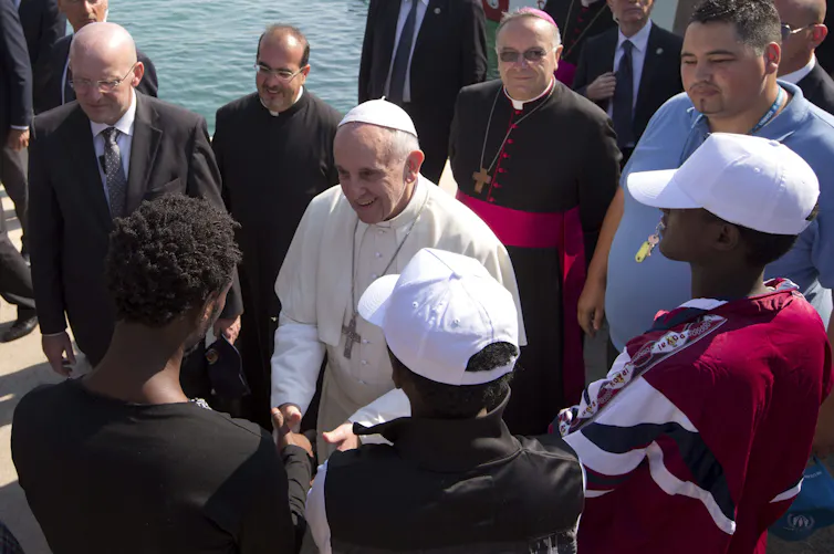 A old man in a white cassock shakes hands with a younger man, other people stand around them.