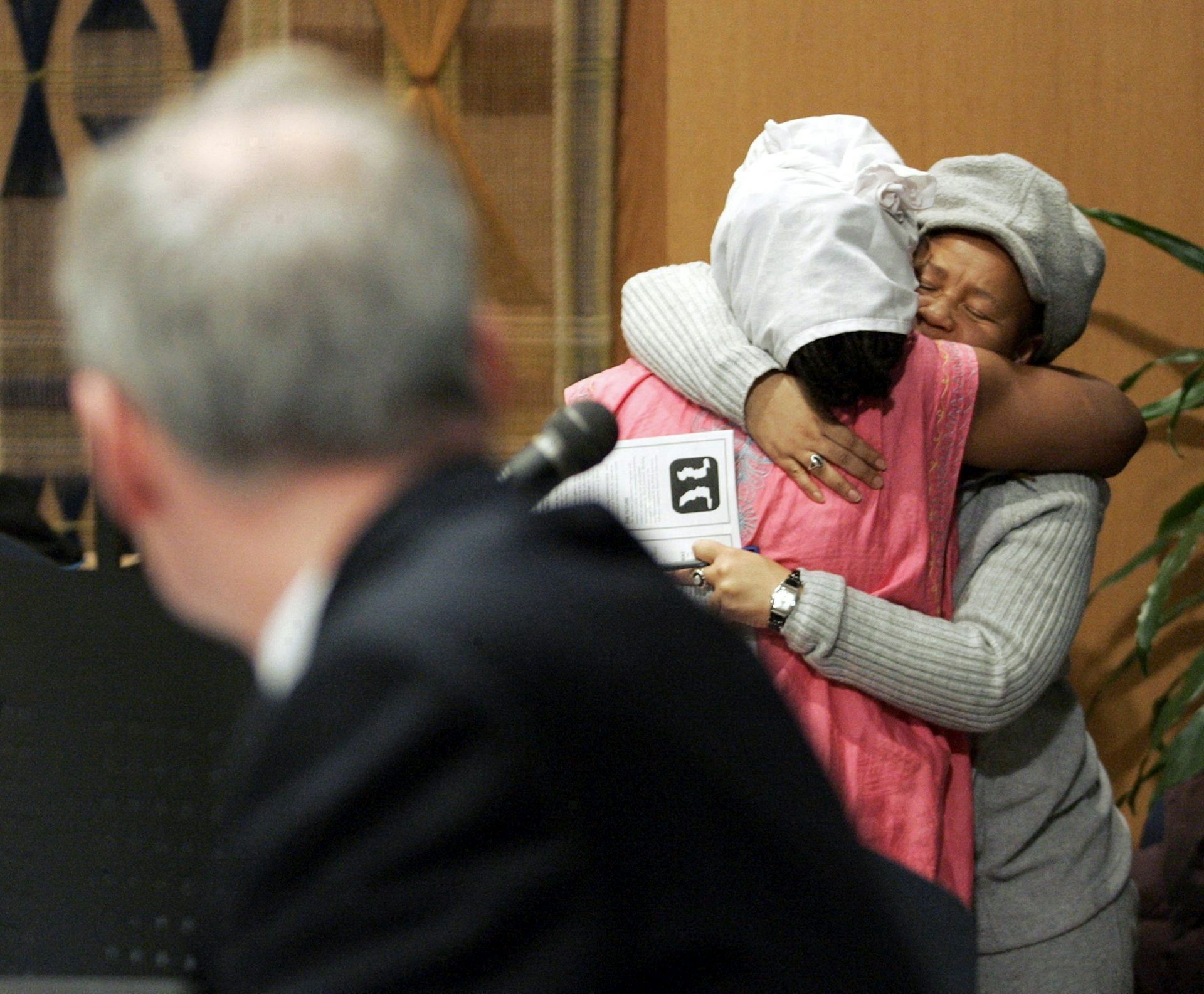 Two Black women seen hugging while a man in suit in fuzzy focus looks over his shoulder.