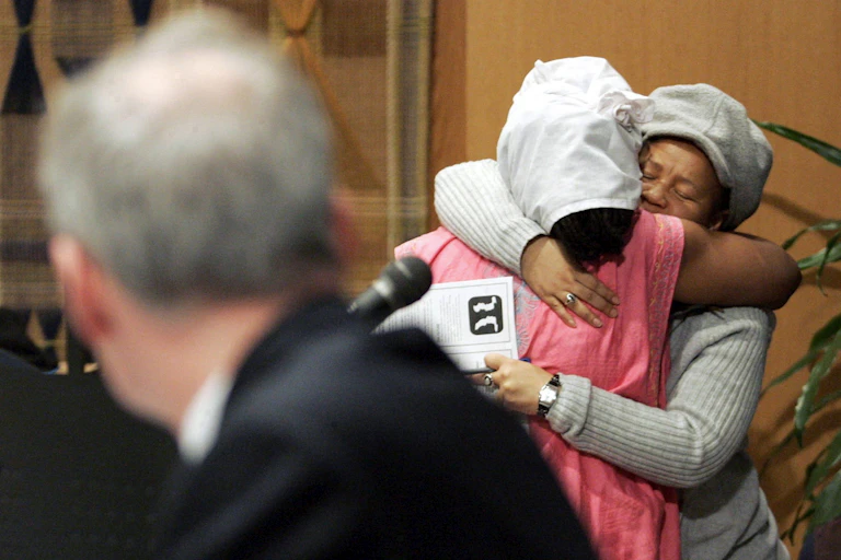 Two Black women seen hugging while a man in suit in fuzzy focus looks over his shoulder.