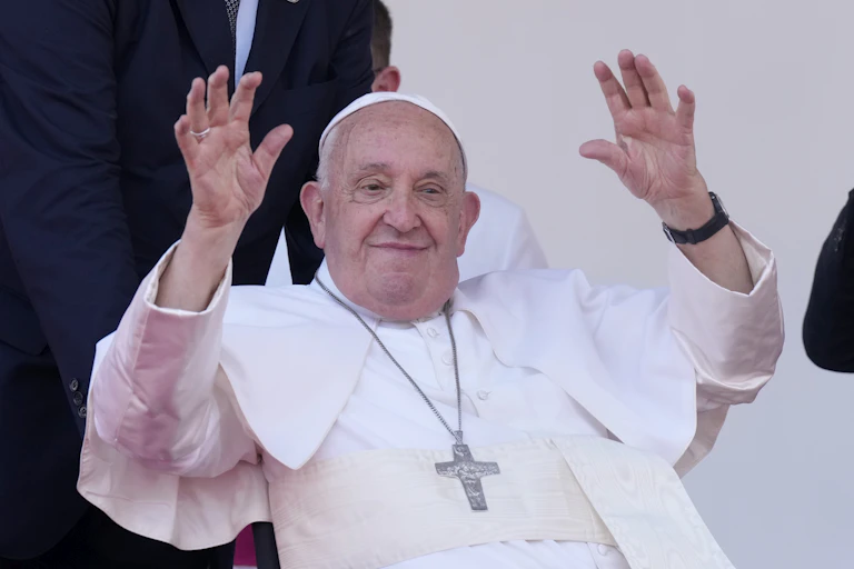 A old man in a white cassock with a silver cross hanging around his neck waves with both hands