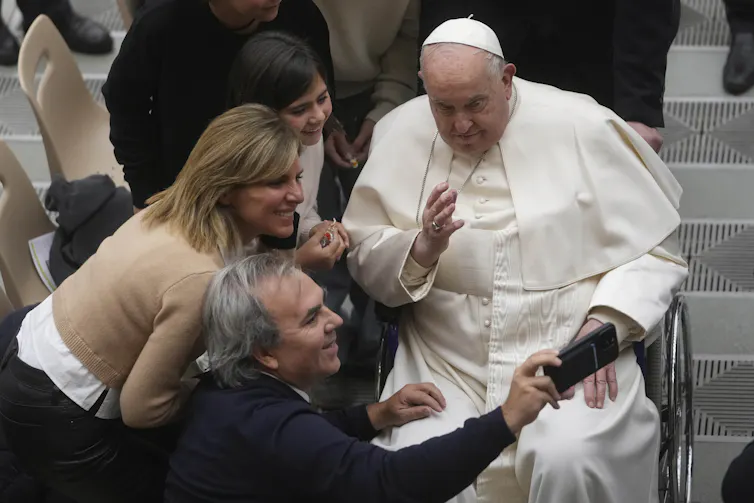 An old man in white cassock sitting in a wheelchair poses for a selfie with a group of people