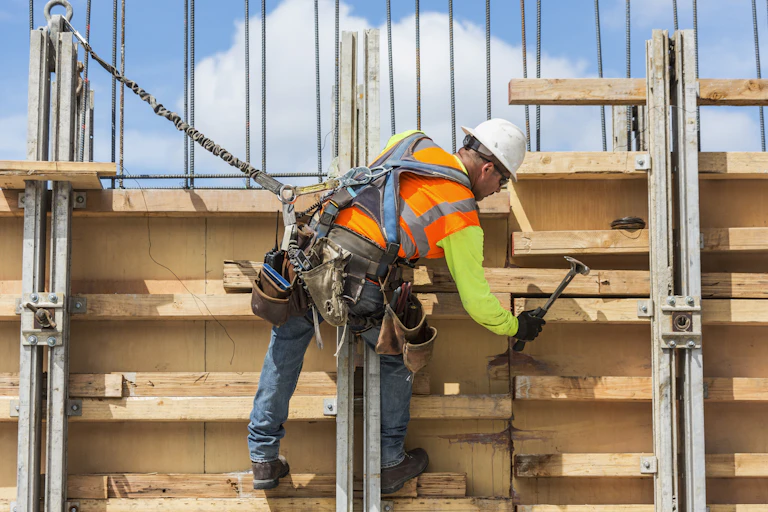 A worker in a reflective vest and hard hat hammers wood at a construction site.