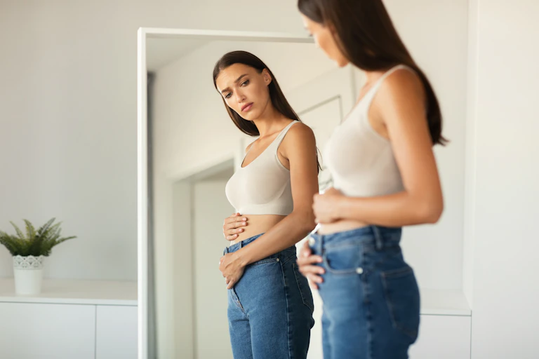 womanwearing blue jeans looks in mirror at her belly