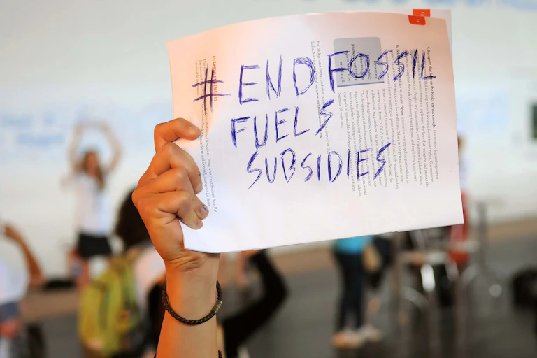 A hand holds up a handwritten sign saying "#End Fossil Fuels Subsidies"