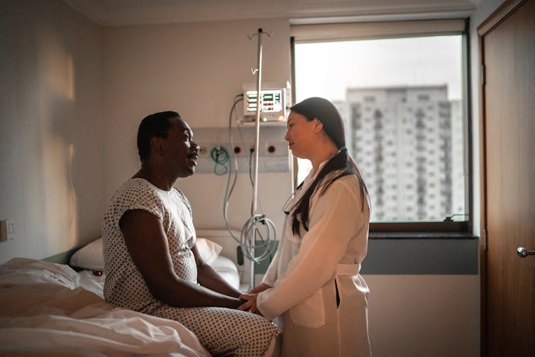 Patient in hospital gown sitting on bed, talking with a doctor in a hospital room