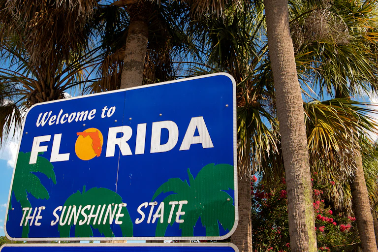 A blue and white sign against palm trees and a blue sky says 'Welcome to Florida, the sunshine state'