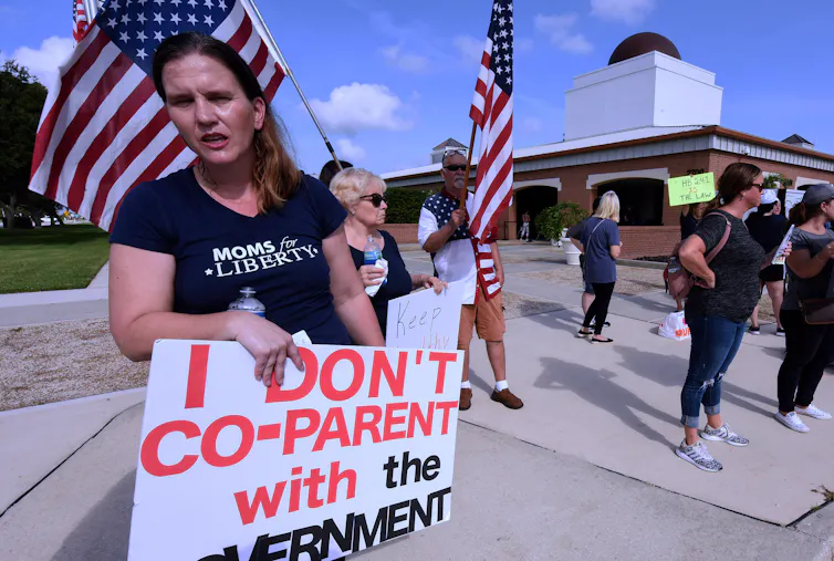 Women stand on a street and hold American flags and a sign that says, 'I don't co-parent with the government.'