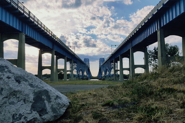 Two parallel blue bridges photographed from low down on the ground between them, looking up to a cloudy sky.