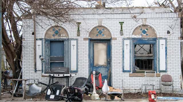 A run-down white brick building is surrounded by trash.