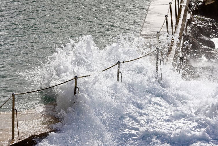 A large wave smashes into an ocean bath through the fencing.