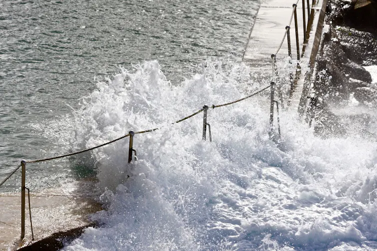 A large wave smashes into an ocean bath through the fencing.