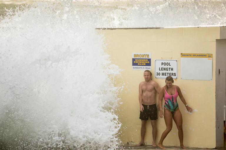 Two swimmers stand on the edge of Bronte baths next to a large frothy wave.