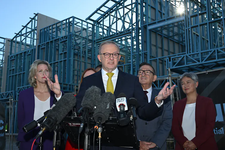 four people stand at microphone in front of housing under construction
