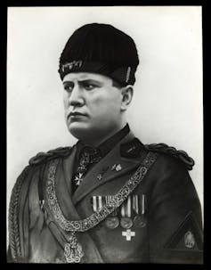 Benito Mussolini wearing military uniform with braid on shoulders, medals and chain with medallion on chest, fur hat.