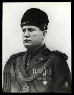 Benito Mussolini wearing military uniform with braid on shoulders, medals and chain with medallion on chest, fur hat.