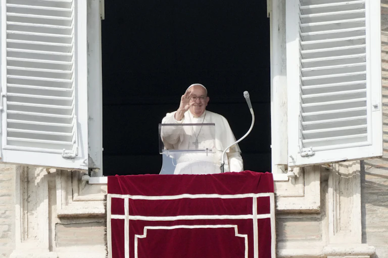 A man in white robes with a large cross around his neck waves from a window, with a microphone in front of him.