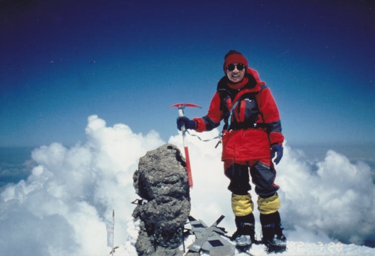 Photo of Tabei in a red coat, standing atop a summit, among the clouds and blue sky