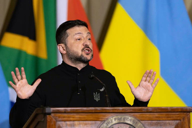 Ukrainian president, Volodymyr Zelensky, holds up both his hands will standing on a stage in front of the Ukrainian and South African flags.