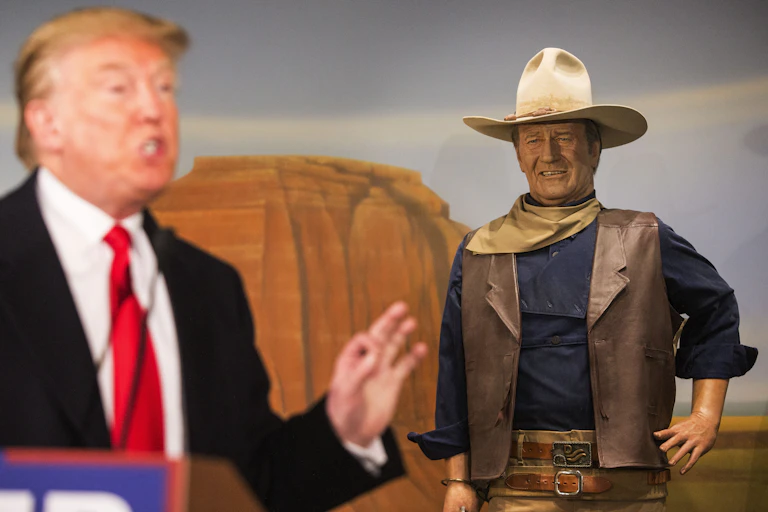 Man in suit gestures to the left of a wax statue of a cowboy.